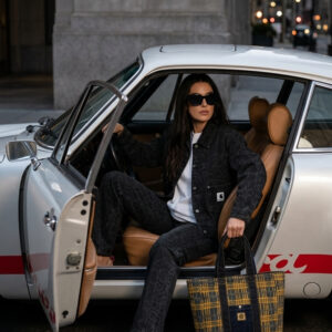 Plan en travelling : Une femme élégante, vêtue d'une tenue en jean noir et de lunettes de soleil, est assise sur le siège passager d'une Porsche 911 Carrera S vintage. La voiture arbore de grands autocollants rouges Carrera sur les bas de caisse et le bas des portières, et son intérieur est en cuir noir. La voiture est garée dans une rue de la ville. La caméra effectue un zoom avant fluide sur elle tandis qu'elle ajuste ses lunettes de soleil, dévoilant son sac cabas tendance sur fond urbain. Elle sort ensuite de la voiture et referme la portière.