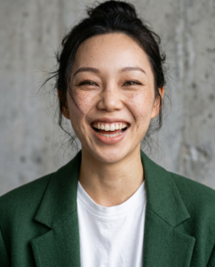 Une femme asiatique de 25 ans aux cheveux châtain foncé, portant un blazer oversize vert de chez Samsøe et un t-shirt oversize blanc, avec un mur de béton en arrière-plan. Éclairage studio zénithal, peau texturée, fond béton froid se trouve à 5m derrière elle donc un peu flou. Portrait Ultra‑détaillé portrait photographique en gros plan, visage d'une femme adulte de 25 ans, peau avec texture réaliste, pores visibles, micro‑imperfections naturelles, subtil dégradé lumineux sur les joues et le front, maquillage léger afin de masquer légèrement les imperfections éclairage doux et flatteur, reflets naturels. Yeux extrêmement détaillés, iris nets, reflets brillants, cils fins et très piqués, mise au point ultra précise sur les cils. Bouche pulpeuse légèrement souriante, contours nets, légère brillance naturelle, rendu réaliste. Elle rigole comme si l'on venait de lui raconter une blague très drôle Style photo haute résolution, prise de vue réalisée avec un boîtier DSLR Sony et un objectif Sigma 80mm f/2. 8,