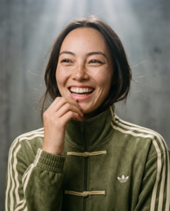Gros plan : Une femme au visage radieux, vêtue d'une veste Adidas verte. La caméra effectue un léger zoom sur son visage souriant, capturant la chaleur de son rire sur un fond texturé à la lumière douce. Soudain, elle éclate de rire, les yeux embués de larmes, à cause d'une blague vraiment nulle qu'on vient de lui raconter. Une seconde auparavant. Puis, d'un geste ample, elle ramène ses cheveux de son front vers l'arrière.