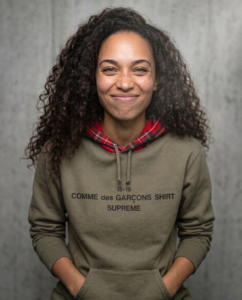 Gros plan : Une femme aux cheveux bouclés sourit chaleureusement, vêtue d'un élégant sweat à capuche à doublure à carreaux. La caméra effectue un léger zoom avant pour capturer son expression joyeuse, mettant en valeur la texture de ses cheveux et les détails de sa tenue sur un fond neutre et doux. Puis elle rit en mettant sa main devant la bouche et en baissant la tête