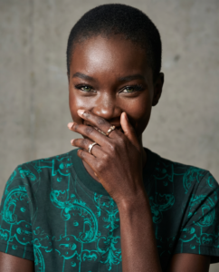 Une femme africaine de 25 ans aux cheveux noirs ultra courts, 3mm, portant un t-shirt vert , avec un mur de croncret en arrière-plan. Éclairage studio zénithal, peau texturée, fond béton froid se trouve à 5m derrière elle donc un peu flou. Portrait Ultra‑détaillé portrait photographique en gros plan, visage d'une femme adulte de 25 ans, peau avec texture réaliste, pores visibles, micro‑imperfections naturelles, subtil dégradé lumineux sur les joues et le front, maquillage léger afin de masquer légèrement les imperfections éclairage doux et flatteur, reflets naturels.